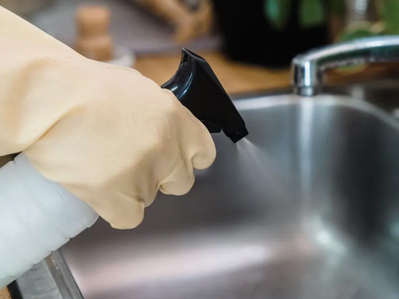 A gloved hand using a spray bottle to clean a stainless steel kitchen sink